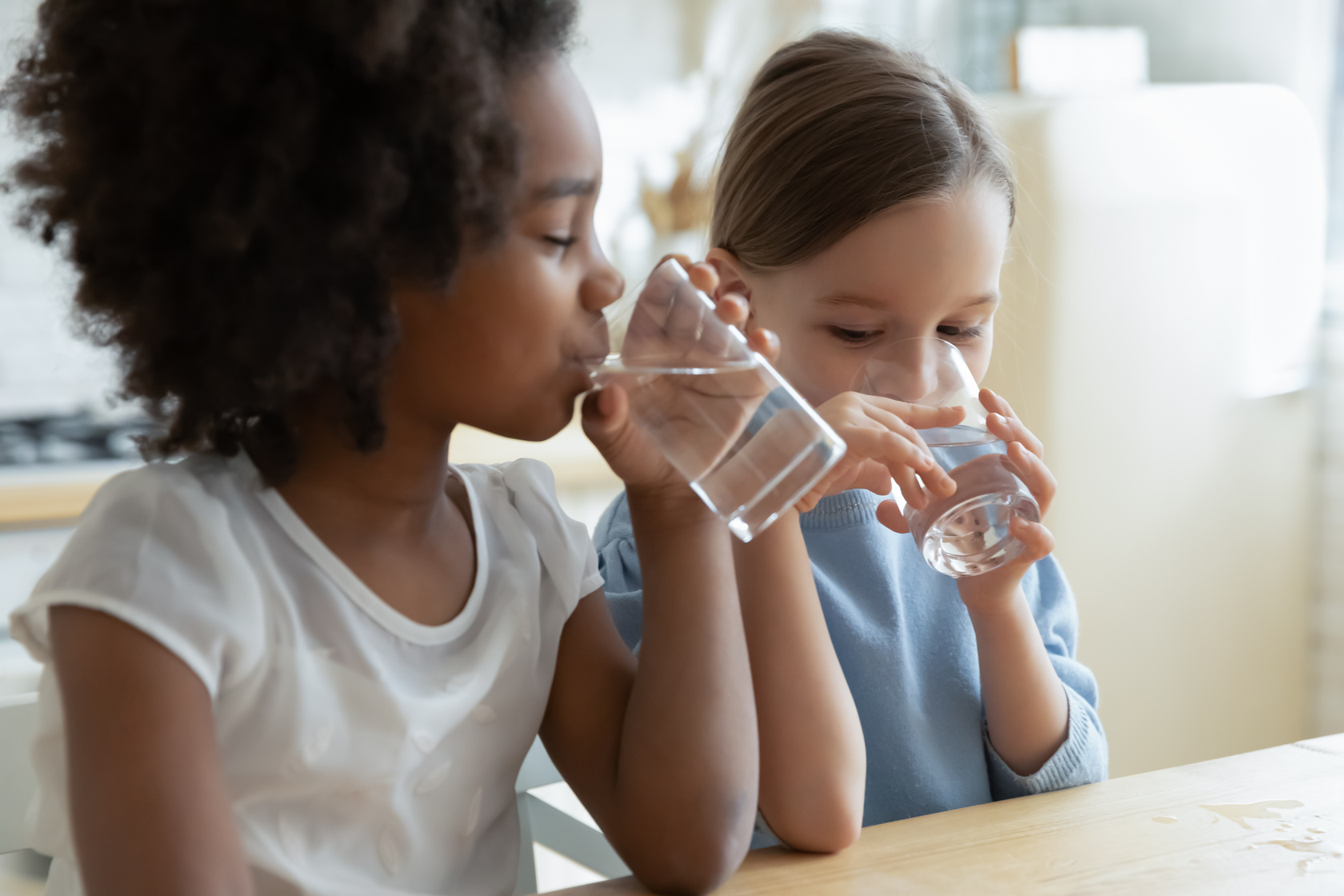 Two multi racial little girls sit at table and drinking filtered water